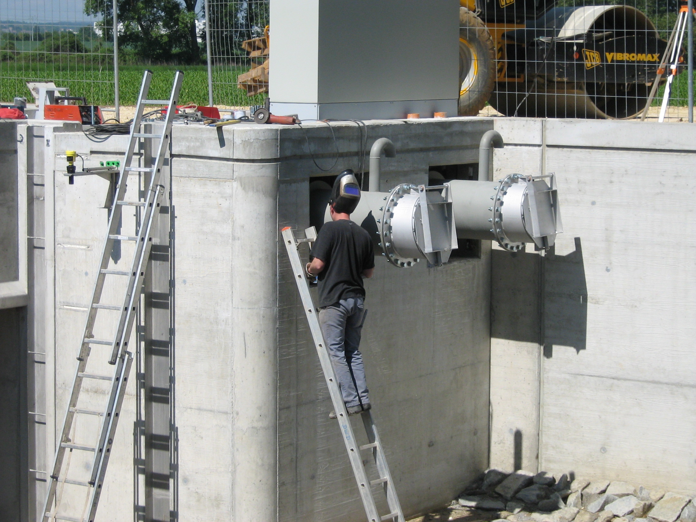 In der Mitte des Bildes lehnt ein Arbeiter mit einer Schweißmaske auf einer Aluminiumleiter an dem Schöpfwerk neben den Rohrschächten. Links davon lehnt eine weitere Aluminiumleiter am betonierten Bauwerk. Oberhalb liegen Werkzeuge, dort steht auch ein Schaltschrank und eine Baumaschine ist im Hintergrund zu erkennen.